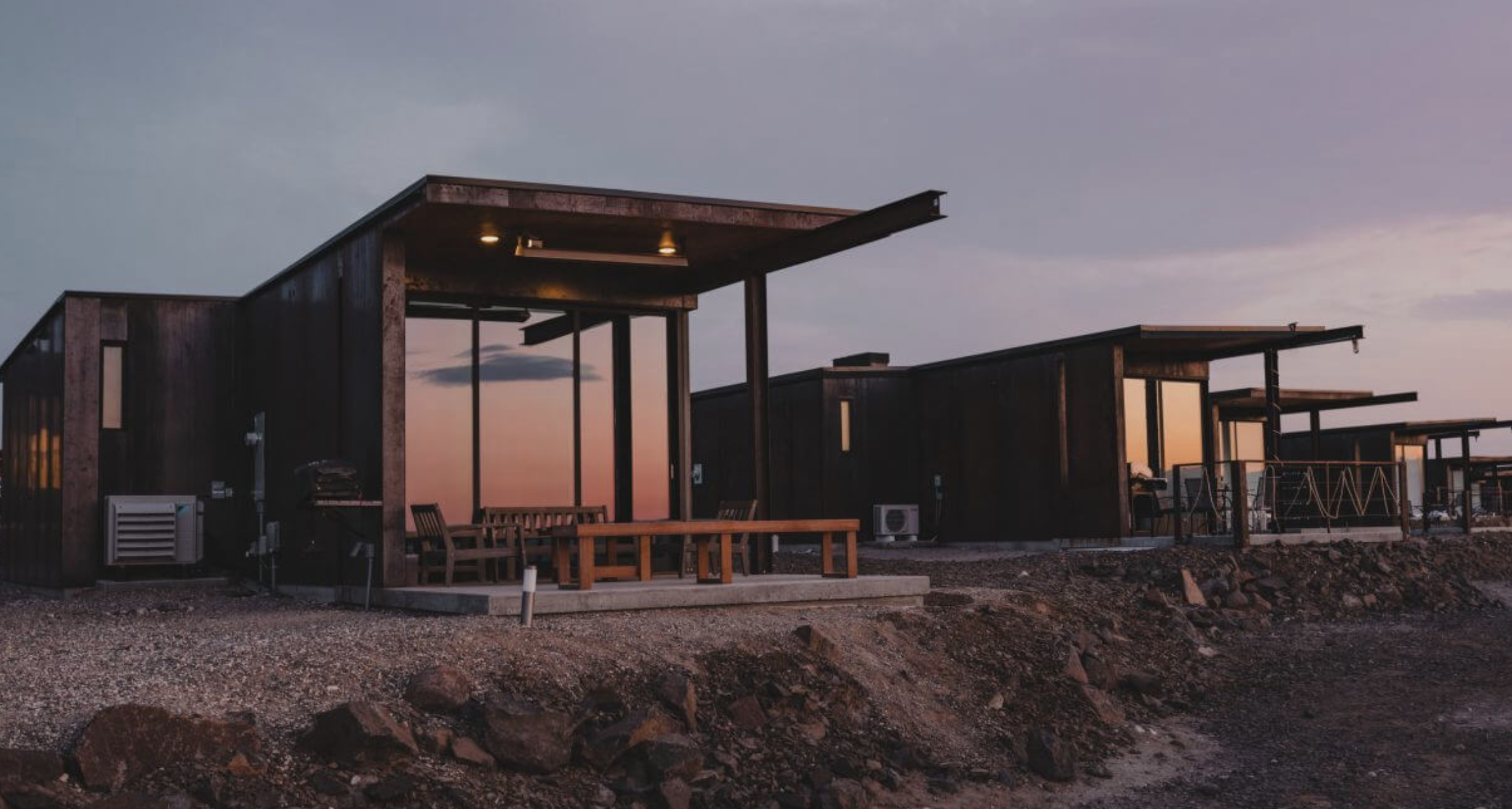 Modern desert-facing villas with minimalist architecture and panoramic glass walls glow in the warm light of sunset, set against a rocky terrain and expansive sky. Sage Outdoor Advisory evaluates high-design glamping accommodations like these in Feasibility Studies to measure visual impact, luxury pricing potential, and guest experience in remote settings.
